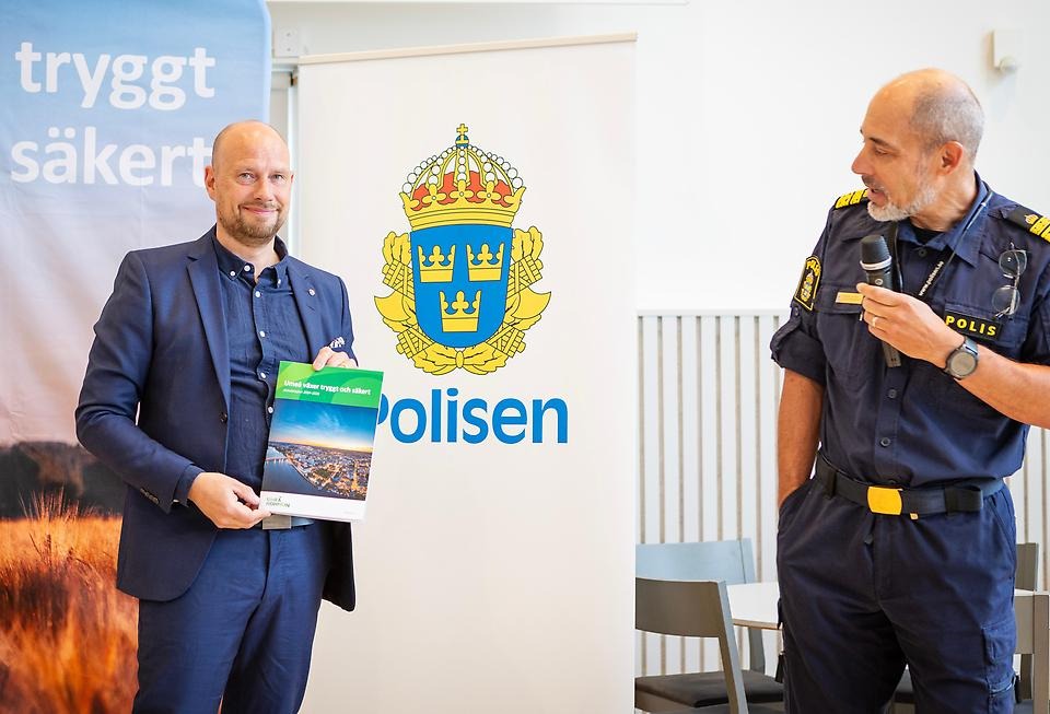 En man håller upp en broschyr tillsammans med en annan man som talar i en mikrofon. 