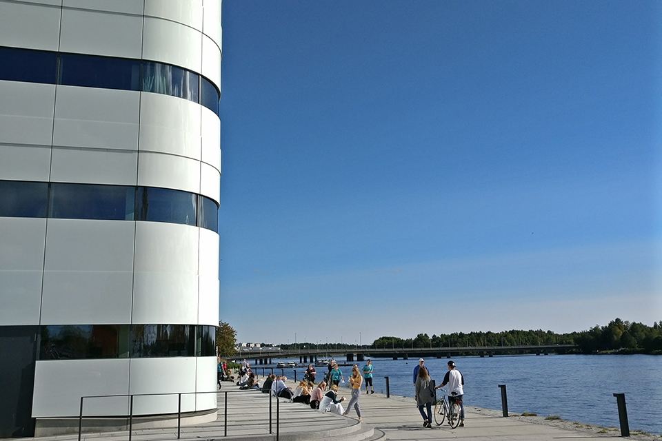 Människor som sitter utanför Väven och promenerar längs strandpromenaden.