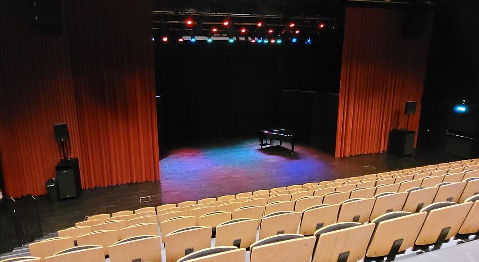 Foto taget på scenen i q-ben. Röda drapperier hänger längs scenen. På scenen står en flygel. Ett stort antal stolsplatser i rad finns framför scenen.