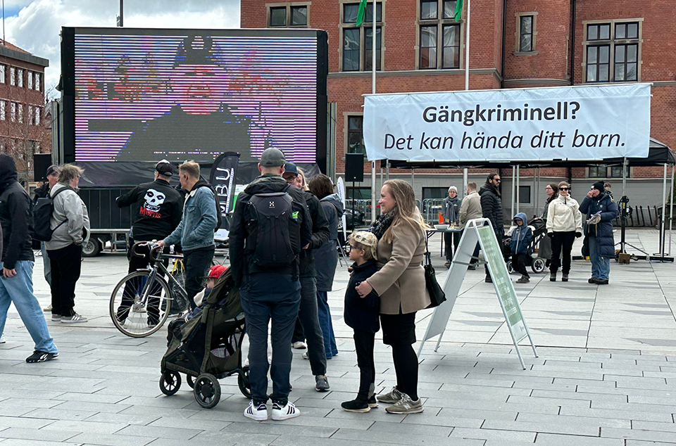 Många människor på torg. Storbildsskärm med polis i bild, gatupratare och vepa med texten "Gängkriminell? Det kan hända ditt barn.".