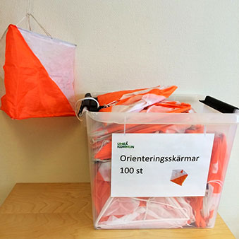 Stor orange och vit orienteringsskärm som hänger upp, många likadana orienteringsskärmar i en låda bredvid.