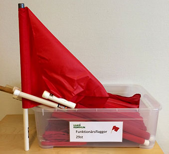 Röd funktionärsflagga med vit flaggpinne, bredvid en låda med fler funktionsflaggor.
