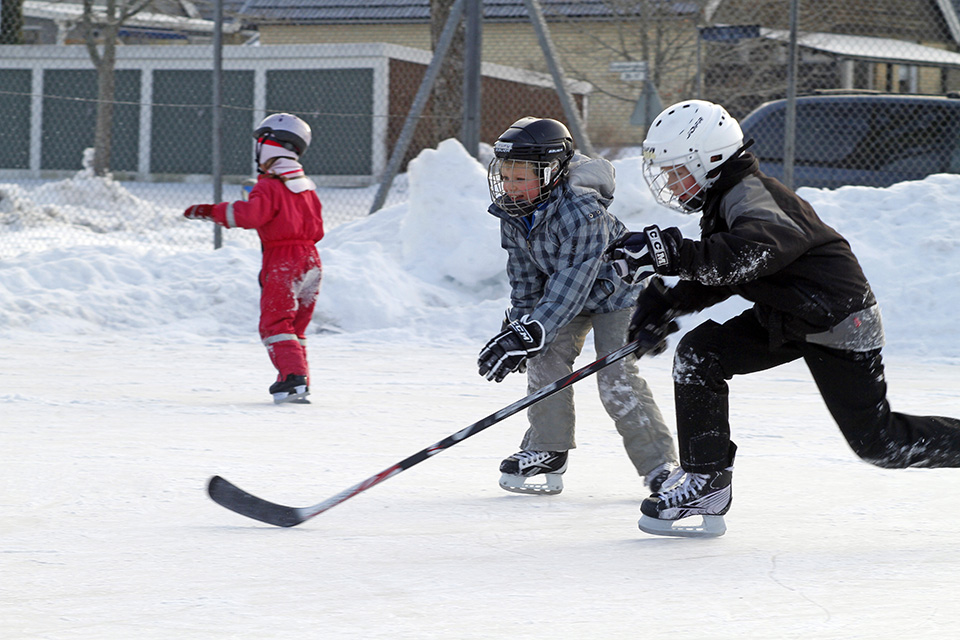 Tre barn leker på isbana. En har en hockeyklubba i handen.