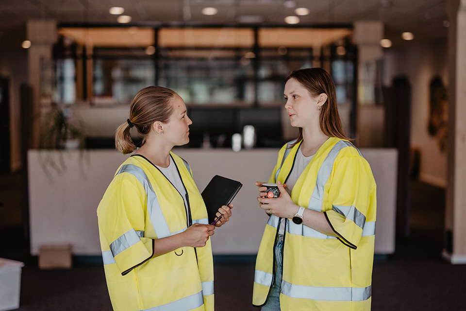 Two women talking. They are dressed as they are working in manufacturing.