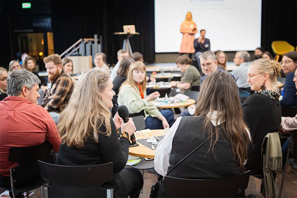 En kvinna sitter med en mikrofon vid ett bord under en workshop och delar sina tankar med gruppen.