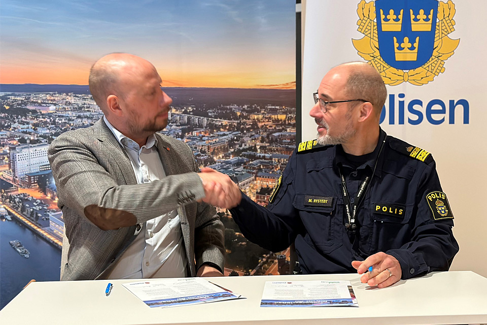 Två män samtalar stående vid ett bord inomhus. Polislogotyp på rollup i bakgrunden.