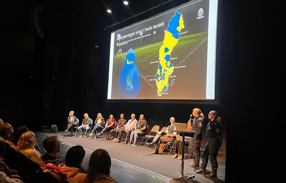 Flera personer sitter på en scen. Två poliser pratar och visar en bild med texten "Rekryteringar sker i hela landet" och en Sverige-karta.
