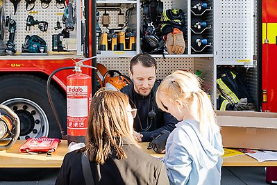Brandman berättar och visar för en kvinna och en flicka, vid en brandbil.