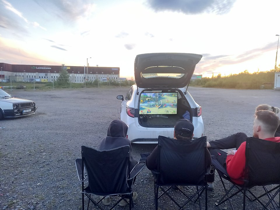Ungdomar utomhus på tältstolar som spelar tv-spel. Tv-spel och skärm finns i bakluckan på en bil.